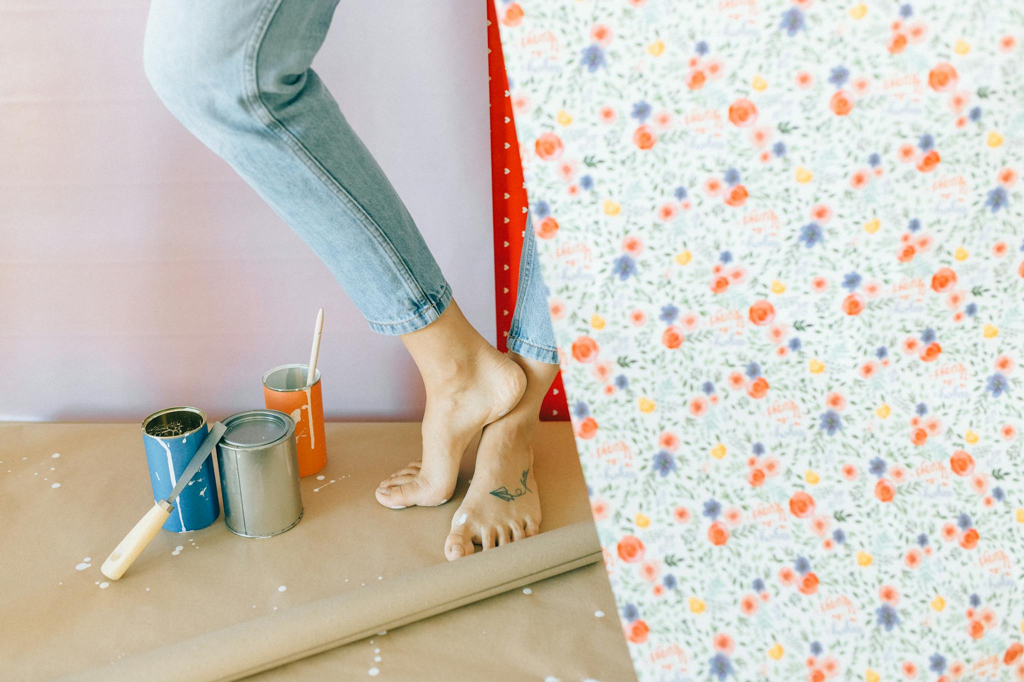 A woman decorates a room with floral wallpaper, surrounded by paint cans and a brush.